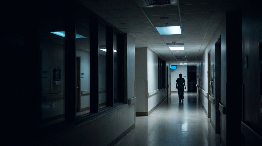 Empty hospital ward corridor at 3am under cold blue-white fluorescent light with a nurse silhouette at the far end
